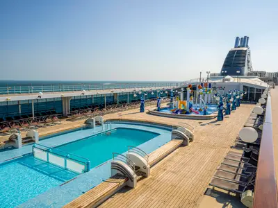 Ein weitreichendes Sonnendeck mit mehreren Pools, Lounge-Stühlen und einem bunten Doremi Spray Park an Bord der MSC Opera. Im Hintergrund ist das blaue Meer zu sehen.