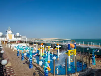 Das Sonnendeck der MSC Opera mit dem bunten Doremi Spray Park und angrenzenden Pools. Im Hintergrund das blaue Meer und eine flache Küstenlinie unter klarem Himmel. Ohne Personen.