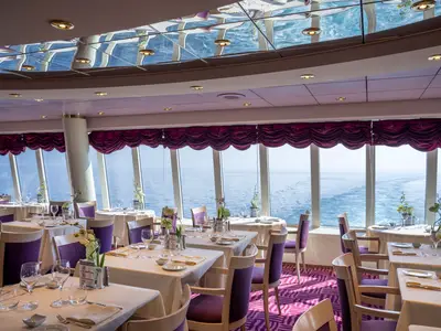 Elegantes Restaurant L'Approdo an Bord der MSC Opera. Tische mit weißen Tischdecken, lila Stühlen und Orchideen. Große Fenster bieten einen weiten Blick auf das tiefblaue Meer und die Heckwelle des Schiffes.