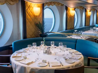 Ein elegantes Restaurant "La Caravella" an Bord der MSC Opera mit weiss gedeckten Tischen und blauen Sitzbänken. Grosse Bullaugen bieten einen weiten Blick auf das offene Meer.