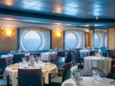 Elegantes Restaurant an Bord der MSC Opera. Gedeckte Tische, Stühle, blaue Sitzbänke und goldene Wandpaneele mit warmen Lichtern. Grosse Bullaugen bieten weiten Meerblick.
