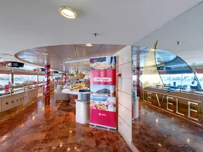 Geräumiges Le Vele Buffetrestaurant der MSC Opera. Helle Buffetheken, viele Tische und Stühle. Grosse Fenster mit Panoramablick auf einen Hafen. Ein rotes 'Bon Voyage' Banner fällt auf.