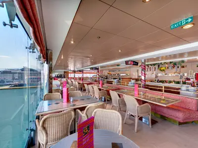Innenansicht des Buffetrestaurants Le Vele an Bord der MSC Opera mit Rattan-Stühlen, gepolsterten Bänken, Buffetinseln und grossen Panoramafenstern mit Blick auf ein anderes Schiff im Hafenbecken.