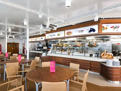 Ein heller Buffetbereich mit mehreren Essensstationen, Tischen und Stühlen an Bord der MSC Opera. Der Fokus liegt auf der Pasta- und Pizza-Station mit Brotkörben und Getränkespendern.