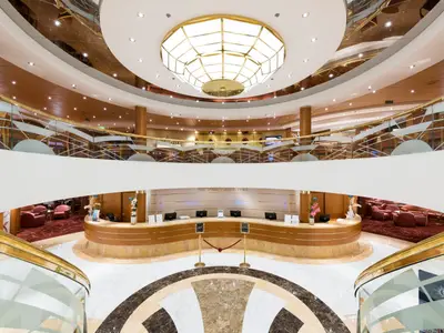 Die elegante Rezeption und das Atrium an Bord der MSC Opera. Geschwungener Empfangstresen, goldene Balustraden und ein prächtiger Kronleuchter dominieren die Szene. Ohne Personen.