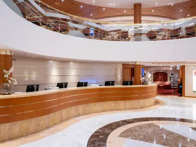 Die Rezeption und der Gästeservice-Schalter an Bord der MSC Opera. Ein geschwungener Tresen mit Marmor- und Holzverkleidung, goldene Akzente und die Aufschrift "Reception - Guest Service". Im Hintergrund ein offenes Atrium mit elegantem Design, Glasbalustraden und einer weiteren Ebene, alles in warmen Brauntönen gehalten. Der Boden ist aus gemustertem Marmor.