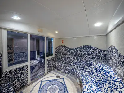 Dampfbad im MSC Aurea Spa der MSC Opera mit geschwungenen, blau-weiss-grauen Mosaikfliesen an Sitzbänken und Wänden. Eine Glastür führt zu einem angrenzenden Wellnessbereich. Helle Deckenbeleuchtung.