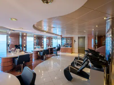 Eine Weitwinkelaufnahme zeigt den luxuriösen Friseursalon an Bord der MSC Opera mit großen Fenstern zum Meer, Styling-Stationen mit beleuchteten Spiegeln und Haarwaschbecken. Der Raum ist in warmen Holztönen und Weiß gehalten, mit modernen schwarzen Stühlen. Keine Personen sind sichtbar.