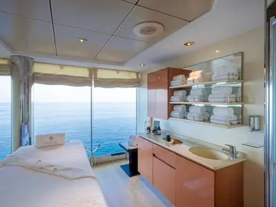 Ein heller Spa-Behandlungsraum an Bord der MSC Opera. Eine Massagebank steht direkt am Fenster mit weitem Meerblick. Regale mit Aurea Spa Handtüchern und ein Waschbecken sind rechts sichtbar.