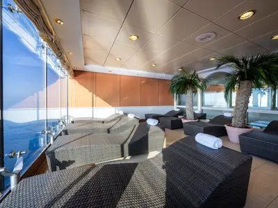 Heller Spa-Ruheraum an Bord der MSC Opera mit Rattan-Liegen, Palmen und großer Glasfront zum Meer. Weiße Handtücher liegen auf den Liegen.