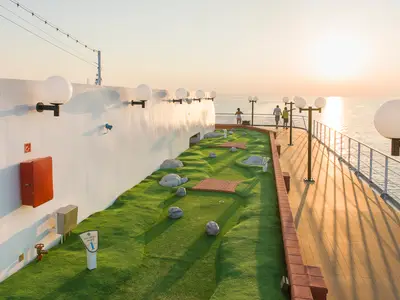 Ein Mini-Golfplatz mit Kunstrasen und Hindernissen auf dem Aussendeck der MSC Opera bei Sonnenuntergang. Im Hintergrund blicken Personen auf das glitzernde Meer.
