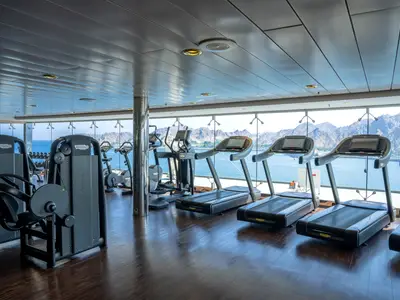 Modernes Fitnessstudio an Bord der MSC Opera mit Technogym-Laufbändern und Kraftgeräten. Panoramablick auf Meer und Berge durch große Fenster.