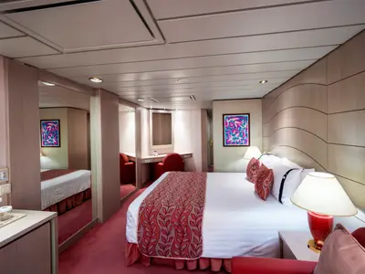 Junior Suite Aurea an Bord der MSC Opera: Großes Doppelbett mit rotem Läufer, Nachttische, Schminkbereich und Spiegel. Helle, moderne Einrichtung in Holz und Weiß mit roten Akzenten.