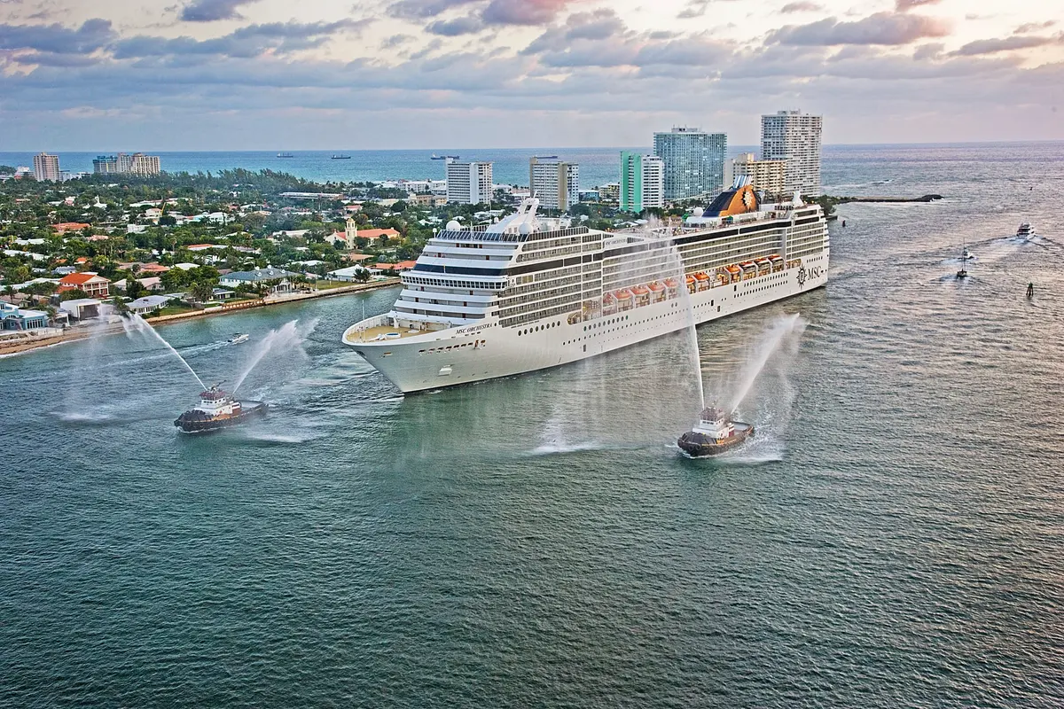 Luftaufnahme der MSC Orchestra, einem großen Kreuzfahrtschiff, das von zwei Schleppern mit Wasserfontänen auf See begleitet wird. Im Hintergrund ist eine Küstenstadt mit vielen Gebäuden zu sehen.