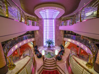 Atrium der MSC Orchestra, aus der Vogelperspektive. Eine lila beleuchtete Wand ist umgeben von modernen Sitzbereichen, Treppen und Glasbalustraden. Das Design wirkt elegant und luxuriös.