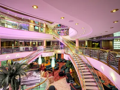 Blick von oben in das weitläufige, elegant beleuchtete Atrium der MSC Orchestra. Eine geschwungene Treppe führt durch mehrere Ebenen mit Sitzbereichen, Palmen und Geschäften.