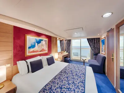 Eine helle Balkonkabine an Bord der MSC Orchestra. Man sieht ein Doppelbett, blauen Teppich, rote Wandakzente, Schreibtisch und einen Balkon mit Meerblick.