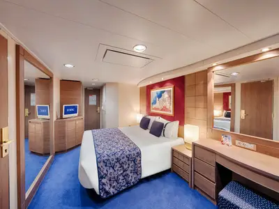 Eine gut beleuchtete Balkonkabine an Bord der MSC Orchestra. Sie zeigt ein komfortables Doppelbett, einen Schreibtisch mit Spiegel und zwei Fernseher. Die Einrichtung ist in Holztönen und blauer Teppich gehalten.