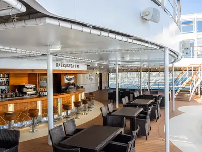 Die helle Barracuda Bar auf dem Außendeck der MSC Orchestra mit Holztheke, Barhockern und Tischen. Im Hintergrund ist ein Hafen mit vielen Yachten und Gebäuden am Horizont zu sehen.