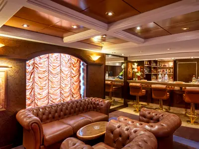 Elegante Bar-Lounge an Bord der MSC Orchestra. Braune Ledersofas, Barhocker und eine bestückte Bar. Warme Beleuchtung und Holzvertäfelung schaffen ein luxuriöses Ambiente.