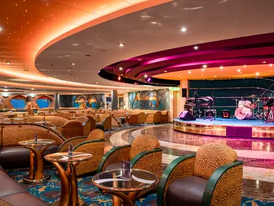 Innenansicht der Savannah Bar der MSC Orchestra. Eine Bühne mit Musikinstrumenten ist rechts. Davor Sitzgruppen mit Leopardenmuster-Sesseln und Tischen. Die Decke ist farbig beleuchtet.