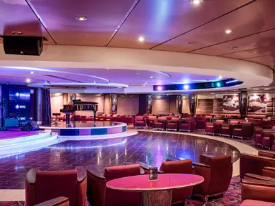 Die Shaker Lounge an Bord der MSC Orchestra. Eine Bühne mit Flügel. Rote Sessel und Tische umgeben die Tanzfläche. Stimmungsvolle violette LED-Beleuchtung.