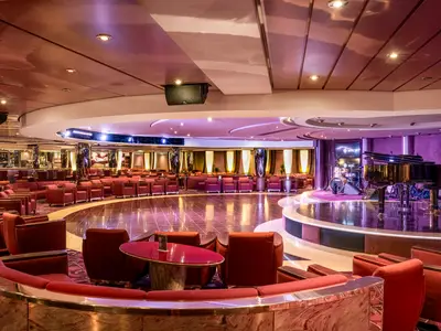 Weitwinkelaufnahme der Shaker Lounge an Bord der MSC Orchestra mit runder Tanzfläche, rotem Mobiliar und Bühne mit schwarzem Flügel und Schlagzeug.