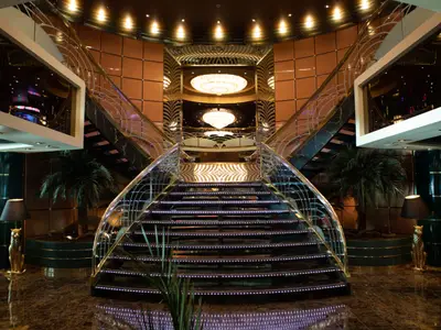 Ein elegantes Treppenhaus an Bord der MSC Orchestra mit schwarzen Marmorstufen und goldener Balustrade. LED-Lichter zieren die Stufen, und zwei prunkvolle Kronleuchter hängen von der Decke. Palmen und goldene Leoparden-Skulpturen schmücken den unteren Bereich.