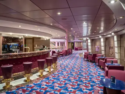Die Zaffiro Bar an Bord der MSC Orchestra, eine stilvolle Lounge mit einer geschwungenen Bar, roten Barhockern und Sesseln sowie einem auffälligen, bunten Teppich.