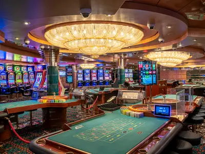 Das Palm Beach Casino an Bord der MSC Orchestra. Reihen leuchtender Spielautomaten und mehrere grüne Roulettetische sind zu sehen. Grosse Kristallleuchter erhellen den Raum und schaffen eine einladende Spielatmosphäre.