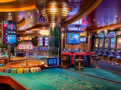 Ein Blick auf das moderne Palm Beach Casino an Bord der MSC Orchestra. Grüne Spieltische mit Roulette und bunten Chips im Vordergrund. Im Hintergrund beleuchtete Spielautomaten und weitere Tische unter warmer Beleuchtung.