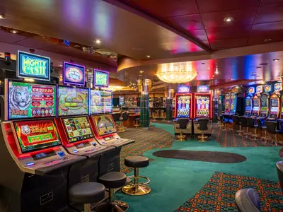 Blick in das Palm Beach Casino an Bord der MSC Orchestra, gefüllt mit Reihen von leuchtenden Spielautomaten und Barhockern auf einem gemusterten Teppich. Die bunte Beleuchtung der Maschinen spiegelt sich in der Decke wider.