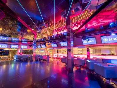 Die R32 Disco an Bord der MSC Orchestra bei Nacht, mit einem leeren Dancefloor, bequemen Sofas und Sesseln, einer beleuchteten Bar und zahlreichen Bildschirmen mit dem MSC Logo. Die Decke ist verspiegelt, und farbige Lichter tauchen den Raum in Magenta- und Blautöne.