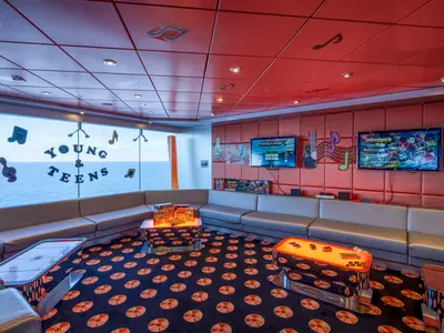 Ein bunter Young & Teen Club an Bord der MSC Orchestra mit gepolsterten Wänden, zwei Flachbildschirmen für Videospiele, bequemen Ledersofas und Tischen. Durch große Fenster ist das Meer sichtbar. Dekorative Musiknoten schmücken den Raum.