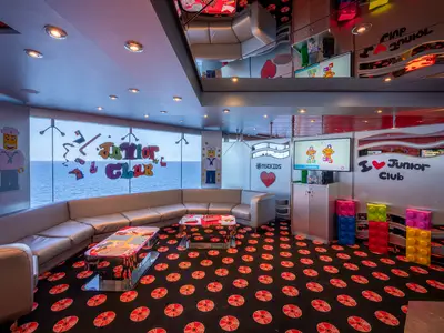 Der farbenfrohe Junior Club an Bord der MSC Orchestra zeigt einen gemütlichen Sitzbereich mit Sofa, Spieltischen, TV-Bildschirmen und Blick auf das Meer. Große Lego-Figuren und Bausteine schmücken den Raum.