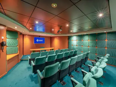 Konferenzraum an Bord der MSC Orchestra. Reihen türkisfarbener Sessel sind auf einen Präsentationstisch und einen Bildschirm mit MSC Logo ausgerichtet. Orange Wände mit grünen Paneelen.