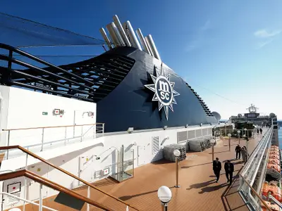 Sonniges Deck der MSC Orchestra mit dem prominenten dunkelblauen Schornstein und MSC-Logo. Mehrere Gäste spazieren auf dem Teakholz-Deck. Im Hintergrund sind das Meer und Hafenanlagen sichtbar.