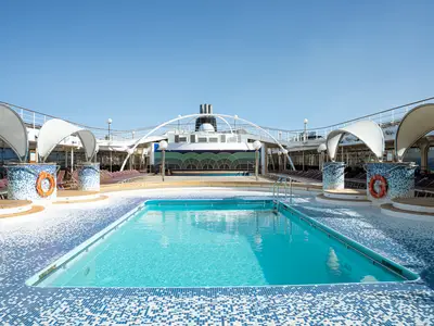 Das sonnige Pooldeck der MSC Orchestra. Ein großer Pool mit blauem Wasser, umgeben von Mosaikfliesen und vielen Liegestühlen. Moderne Sonnensegel und der Schiffsaufbau mit Schornsteinen sind ebenfalls sichtbar.