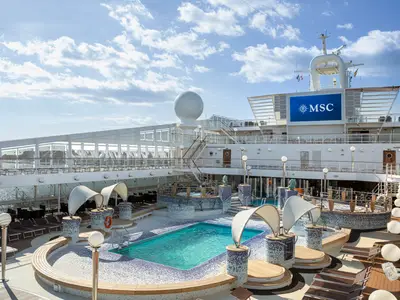 Detailansicht des weitläufigen Poolbereichs an Bord der MSC Orchestra mit leuchtend blauem Wasser, vielen Liegestühlen und einem großen MSC-Bildschirm im Hintergrund. Sonnenlicht fällt auf das Deck.