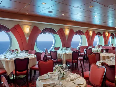 Elegantes Restaurant Villa Borghese auf der MSC Orchestra. Rote Stühle, gedeckte Tische und Meerblick durch Bullaugen. Roter Teppich, Samtpaneele und Wandlampen.