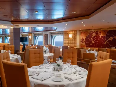 Restaurant L'Ibiscus an Bord der MSC Orchestra mit gedeckten Tischen, orangen Stühlen und Bullaugen zum Meer. Eine florale Wandkunst ziert den Raum.