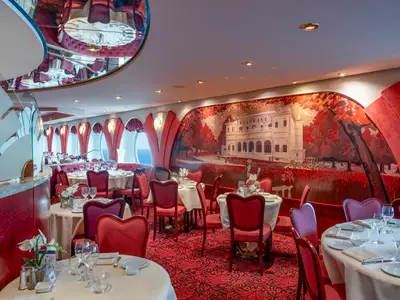 Das elegante Villa Borghese Restaurant an Bord der MSC Orchestra. Rote Sessel, weiß gedeckte Tische und eine Wandmalerei mit einer Villa und roten Bäumen. Runde Bullaugen bieten Meerblick.