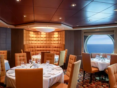 Elegantes Restaurant L'Ibiscus der MSC Orchestra. Gedeckte Tische, orange Stühle, gepolsterte Sitzbank. Bullauge zeigt weiten Blick auf das blaue Meer.