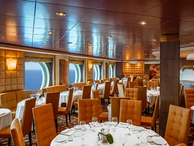 Ein stilvolles Restaurant an Bord der MSC Orchestra. Die Tische sind gedeckt, orangefarbene Stühle und Polster prägen den Raum. Durch Bullaugen ist das Meer sichtbar. Die Decke reflektiert Licht.