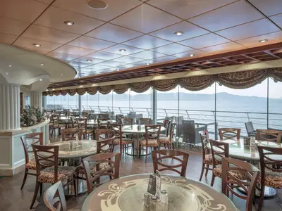 Blick in das Buffetrestaurant La Piazzetta der MSC Orchestra. Viele Tische, Stühle und grosse Fenster mit weitem Meerblick auf Berge. Holzgetäfelte Decke mit Einbauspots.