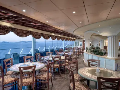 Helles Buffetrestaurant an Bord der MSC Orchestra. Tische und Stuehle stehen an grossen Panoramafenstern mit weitem Meerblick. Saeulen und Gruenpflanzen zieren den Innenraum.