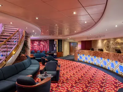 Empfangsbereich an Bord der MSC Orchestra. Geschwungene Rezeption, links eine Wendeltreppe. Dunkelblaue Lounge-Möbel auf rotem Teppich.