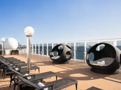 Das Sonnendeck der MSC Orchestra mit vielen schwarzen Liegestühlen und zwei runden Lounge-Sesseln mit Kissen. Eine Glasbrüstung gibt den Blick auf das blaue Meer und den Himmel frei. Holzfußboden und moderne Laternen.