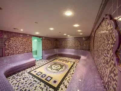 Ein Thermalbereich im MSC Aurea Spa der MSC Orchestra mit lila Mosaikfliesen an Wänden und geschwungenen Bänken. Ein kunstvolles Bodenmosaik ist in der Mitte zu sehen. Im Hintergrund ein Duschbereich.
