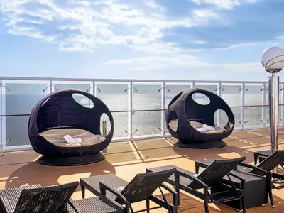 Zwei moderne Lounge-Sessel und mehrere Sonnenliegen stehen auf dem Teakholz-Sonnendeck der MSC Orchestra. Im Hintergrund glitzert das Meer hinter einer Glasbalustrade unter blauem Himmel. Handtücher liegen auf den Sesseln bereit.